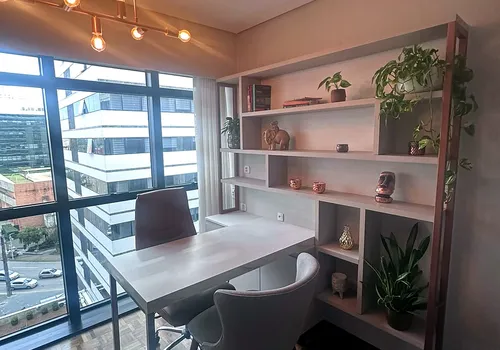 Cantinho de trabalho com estante iluminada, plantas e luminária pendente, no terceiro consultório